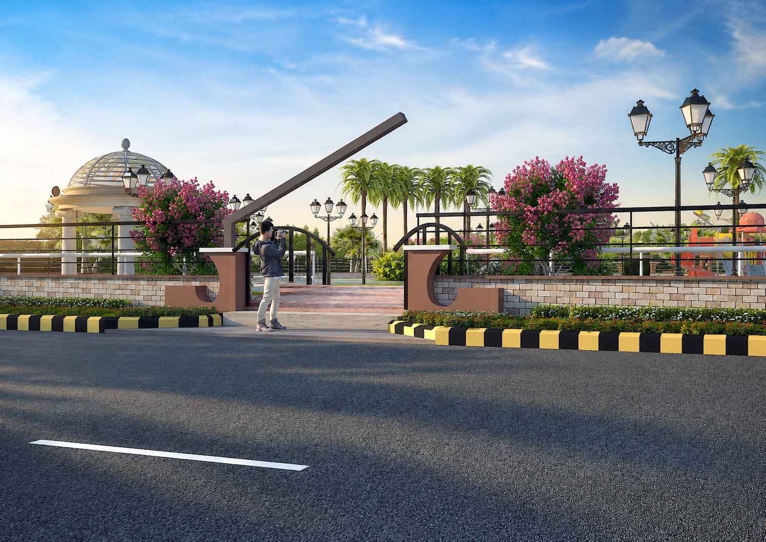 Landscaped entrance gate of a community park with a boom barrier, decorative pillars, flowering plants, and a person taking a photo at the gate.